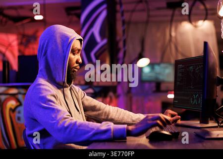 Serveur de piratage criminel d'Internet, craquage de mot de passe et vol d'informations. Homme afro-américain dans la planification de la fraude en ligne de hotte et de codage ransomware sur l'ordinateur dans la chambre noire Banque D'Images