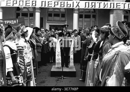 Sloviansk, années 1980, place de la gare. Une captivante photographie d'archives en noir et blanc représentant un travailleur ferroviaire vétéran décoré en uniforme officiel prononçant un discours inspirant devant un microphone. L'orateur se tient sur la scène près du bâtiment du chemin de fer de Donetsk, entouré de jeunes femmes en costumes nationaux ukrainiens traditionnels tapant avec enthousiasme les mains. La foule rassemblée honore le mouvement syndical Pyotr Krivonos, mettant en valeur une profonde fierté industrielle et une forte unité communautaire dans le Donbass pacifique. Banque D'Images