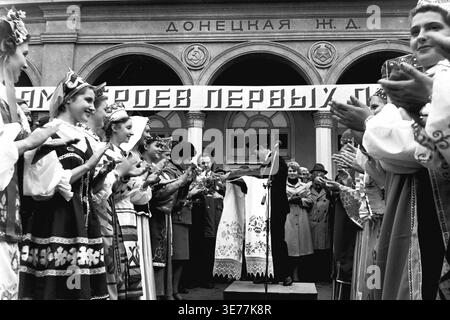 Sloviansk, années 1980, place de la gare. Une captivante photographie d'archives en noir et blanc représentant un travailleur ferroviaire vétéran décoré en uniforme officiel prononçant un discours inspirant devant un microphone. L'orateur se tient sur la scène près du bâtiment du chemin de fer de Donetsk, entouré de jeunes femmes en costumes nationaux ukrainiens traditionnels tapant avec enthousiasme les mains. La foule rassemblée honore le mouvement syndical Pyotr Krivonos, mettant en valeur une profonde fierté industrielle et une forte unité communautaire dans le Donbass pacifique. Banque D'Images