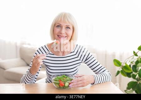 Heureuse dame senior mangeant de la salade fraîche à la maison Banque D'Images