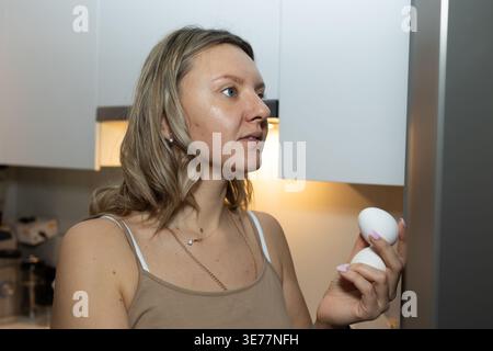 Femme tenant deux œufs frais dans la cuisine moderne près du réfrigérateur. Préparation du petit déjeuner matinal et concept de routine alimentaire saine pour la nutrition quotidienne et les repas de famille. Banque D'Images