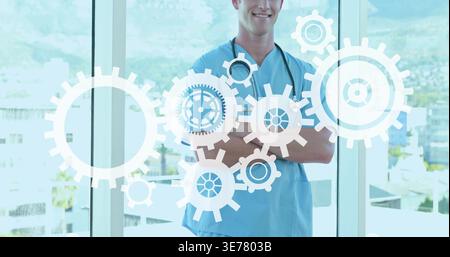 Médecin debout portant des gommages bleu clair et stéthoscope aux fenêtres de la clinique, avec recouvrement de rouage Banque D'Images