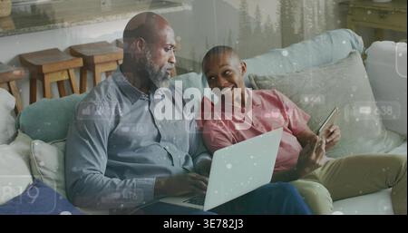 Père afro-américain et fils adolescents assis sur un canapé modulable de couleur claire tenant un ordinateur portable et un ph Banque D'Images