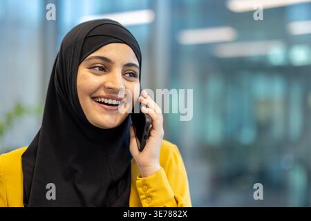 Femme musulmane en hijab et chemise jaune ayant une conversation joyeuse sur son téléphone portable, communiquant activement tout en travaillant dans un bureau moderne envir Banque D'Images