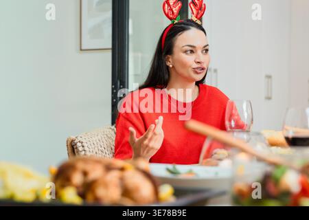 Femme mi-adulte assise à la table à manger en pull rouge et bandeau de coeur, parlant au-dessus de la turquie. Festif, conversation, rassemblement, maison, verrerie, di Banque D'Images