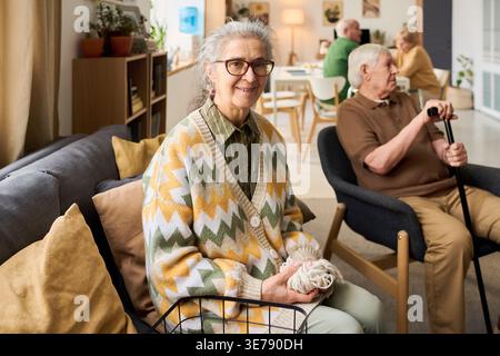 Portrait d'une femme caucasienne senior tricotant dans le salon de la maison de retraite, souriant à la caméra tandis que des résidents âgés socialisent à proximité. Idéal pour les soins aux aînés, l'assistance à la vie, le marketing des services vieillissants Banque D'Images