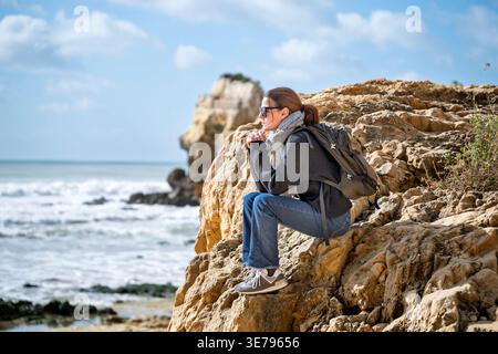 femme est assise sur une falaise côtière rocheuse avec un sac à dos regardant vers la mer Banque D'Images