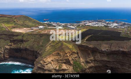 La vue aérienne de Madère montre des falaises volcaniques, une petite crique et l'Atlantique. Panneaux solaires et réservoirs alignés vers le port de Canical avec brise-lames, Banque D'Images