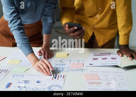 Homme noir tenant smLifestylephone avec une femme caucasienne tenant un stylo pointant sur le document tout en se tenant les mains penchées à la table dans le bureau Banque D'Images