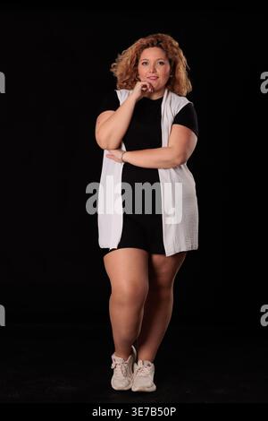 Femme hispanique de grande taille avec des cheveux bouclés blonds touchant le menton debout devant un fond noir en studio Banque D'Images