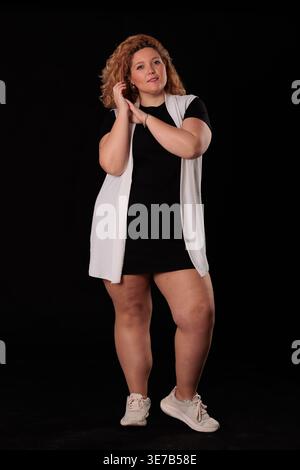 Femme hispanique de grande taille avec les cheveux bouclés debout avec les mains ensemble devant un fond noir en studio Banque D'Images