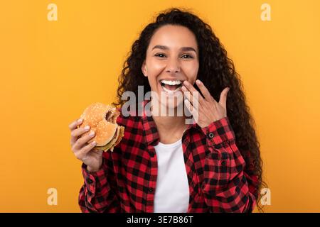Femme joyeuse du moyen-Orient avec les cheveux bouclés mangeant hamburger riant et essuyant les lèvres avec le doigt tout en se tenant sur fond jaune dans le studio Banque D'Images