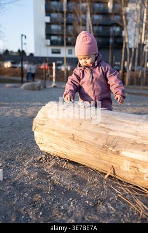 portrait franc ensoleillé d'un tout-petit curieux dans un bonnet rose et une veste violette explorant une grande bûche altérée dans une cour de parc urbain à golden ho Banque D'Images