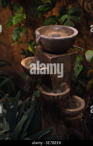 fontaine d'eau en pierre à étages dans un cadre de jardin intérieur luxuriant, bruns terreux chauds et verts profonds, eau en cascade sur des bols texturés et des socles, donc Banque D'Images