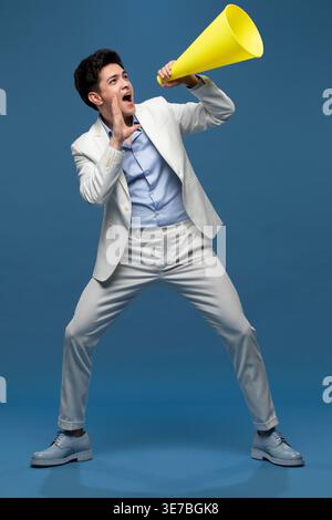 Homme chinois criant avec mégaphone debout avec les jambes écartées sur fond bleu dans le studio Banque D'Images