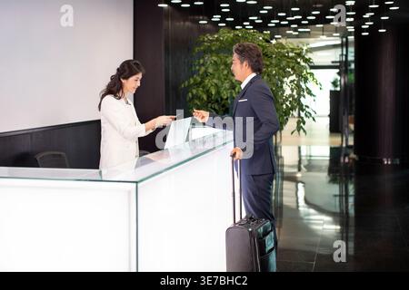 Souriant homme d'affaires chinois d'âge moyen avec des bagages payant son hébergement avec carte de crédit à la réceptionniste chinoise tout en se tenant debout à... Banque D'Images