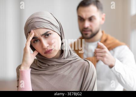 Femme musulmane arabe déprimée en hijab touchant sa tête alors qu'elle se faisait crier dessus par son mari caucasien à la maison Banque D'Images