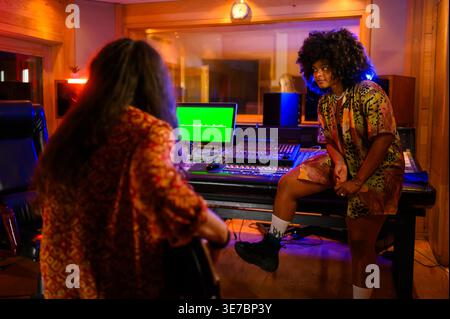 Musicienne hispanique sérieuse avec afro regardant loin assis à côté d'une console de mixage avec lumière néon écoutant l'enregistrement de la lecture de guitare... Banque D'Images