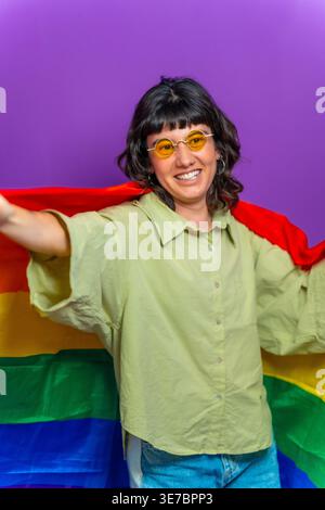 Femme hispanique souriante portant des lunettes de soleil enveloppées d'un drapeau de fierté LGBTQ tout en dansant sur fond violet en studio Banque D'Images