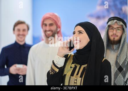 Femme d'affaires joyeuse du moyen-Orient dans des vêtements musulmans traditionnels ayant un appel téléphonique debout avec des collègues masculins sur le lieu de travail Banque D'Images