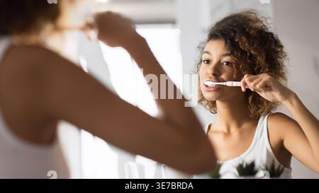 Femme noire heureuse tenant brosse à dents nettoyant les dents devant le miroir à la salle de bain Banque D'Images