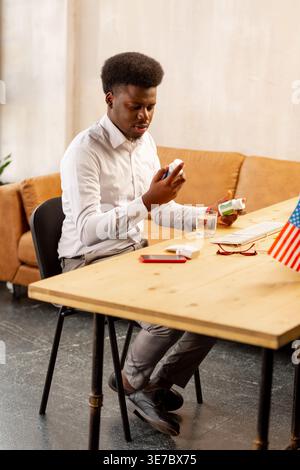 Jeune homme noir regardant des pilules assis à table près du canapé dans le bureau Banque D'Images
