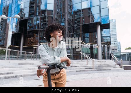 Femme hispanique souriante avec afro bouclé regardant loin se penchant sur le scooter électrique en ville pendant la journée Banque D'Images