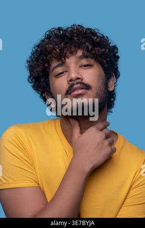 Homme indien sérieux avec les cheveux bouclés debout avec la main sur le cou sur fond bleu Banque D'Images