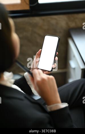 Femme d'affaires asiatique tenant le stylo à l'aide smLifestylephone tout en étant assis à la table dans le bureau Banque D'Images