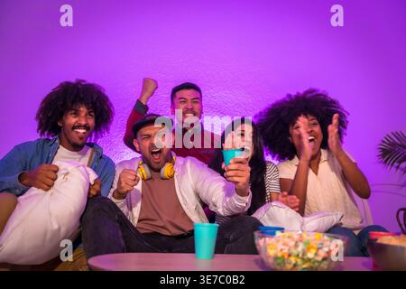 Joyeux groupe diversifié d'amis regardant le jeu intense à la télévision célébrant tout en étant assis ensemble sur le canapé près de la table avec pop-corn et soda dans la vie... Banque D'Images