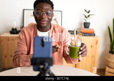 Homme noir souriant faisant du streaming en direct montrant du jus vert sain assis à table à la maison Banque D'Images