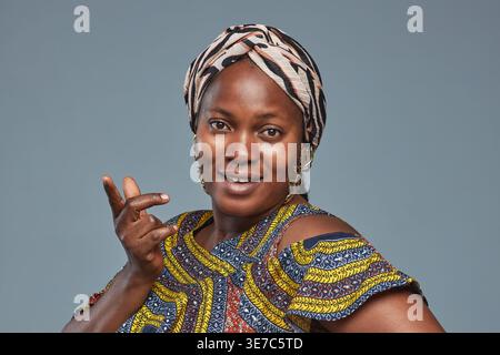 Femme noire portant une robe traditionnelle et un couvre-tête faisant des gestes de la main tout en se tenant debout sur fond gris dans le studio Banque D'Images