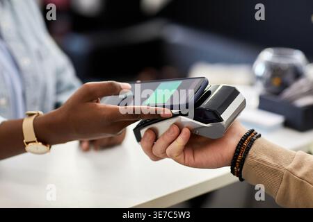 Mains d'une femme noire payant à l'aide d'un smartphone à un caissier masculin caucasien tenant un terminal POS tout en se tenant debout au-dessus du comptoir dans un magasin de vêtements Banque D'Images