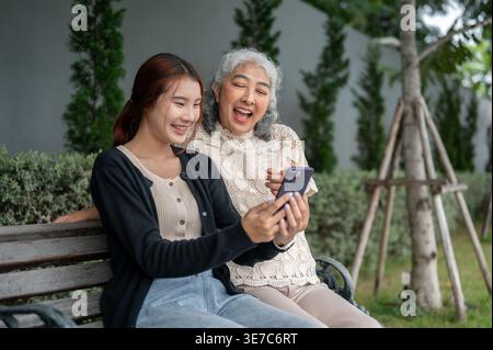 Femme thaïlandaise ayant du plaisir à partager smartphone avec sa grand-mère tout en étant assise sur un banc en bois dans le jardin pendant la journée Banque D'Images