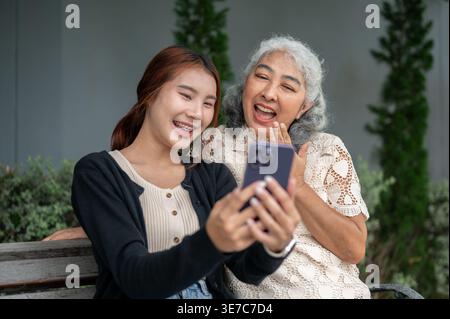 Femme thaïlandaise ayant du plaisir à partager smartphone avec sa grand-mère tout en étant assise sur un banc en bois dans le jardin pendant la journée Banque D'Images