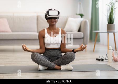 Femme noire avec des vêtements de sport bouclés et casque VR méditant sur tapis dans le salon à la maison Banque D'Images