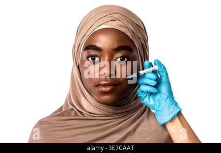 Femme musulmane noire sérieuse portant le hijab recevant un traitement sur le visage du médecin tout en se tenant devant un fond blanc dans le studio Banque D'Images