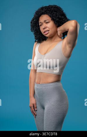 Femme noire avec des douleurs au cou bouclées tout en se tenant sur fond bleu en studio Banque D'Images