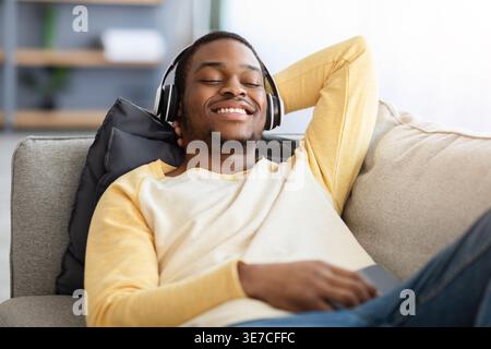 Homme noir souriant couché sur le canapé écouter de la musique avec des écouteurs dans le salon à la maison Banque D'Images