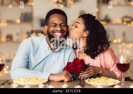Homme noir avec la barbe donnant bouquet de roses rouges à la petite amie l'embrassant sur la joue tout en étant assis à table avec de la nourriture et des boissons célébrant valenti... Banque D'Images
