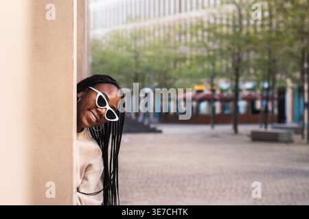 Femme noire avec des tresses portant des lunettes de soleil debout et jetant un coup d'œil hors du pilier du bâtiment dans la ville pendant la journée Banque D'Images