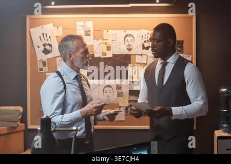 Détectives noirs et caucasiens tenant des notes discutant tout en se tenant debout à côté des preuves à bord dans le bureau Banque D'Images