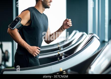 Image recadrée d'homme noir avec la barbe portant le brassard regardant loin jogging sur le tapis roulant dans la salle de gym Banque D'Images