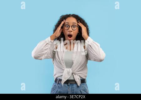 Adolescente noire portant des lunettes avec une expression choquée touchant la tête tout en se tenant debout avec sac à dos sur fond bleu en studio Banque D'Images