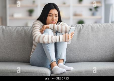 Femme asiatique triste utilisant un smartphone serrant les genoux tout en étant assis sur le canapé dans le salon à la maison Banque D'Images