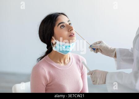 Femme indienne malade portant un masque facial se faisant écouvillonner par le médecin tout en étant assise sur la chaise dans la clinique Banque D'Images