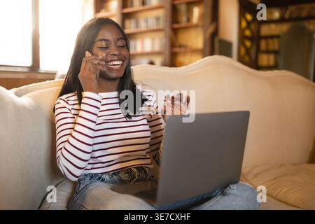 Adolescente noire ayant un appel téléphonique assis avec un ordinateur portable sur le canapé dans le salon à la maison Banque D'Images