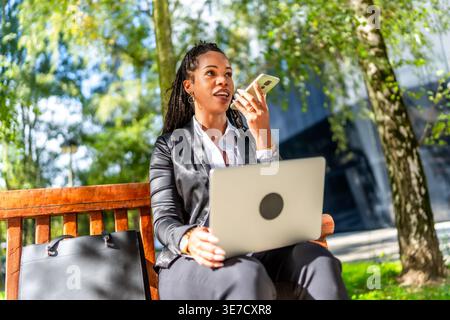 Femme d'affaires hispanique enregistrant un message vocal en utilisant smLifestylephone regardant loin assis avec ordinateur portable sur banc en bois dans le parc le jour ensoleillé Banque D'Images