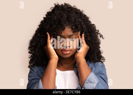 Femme noire avec les yeux fermés touchant le temple tout en se tenant debout sur fond beige dans le studio Banque D'Images