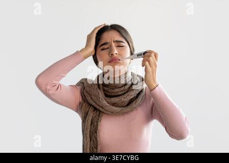 Femelle indienne malade portant une écharpe tenant la tête avec une main vérifiant la température à l'aide d'un thermomètre debout avec les yeux fermés devant le dos gris... Banque D'Images